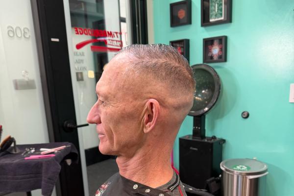 Senior man with a fresh fade haircut, seated in barber chair wearing a black cape, teal wall in background