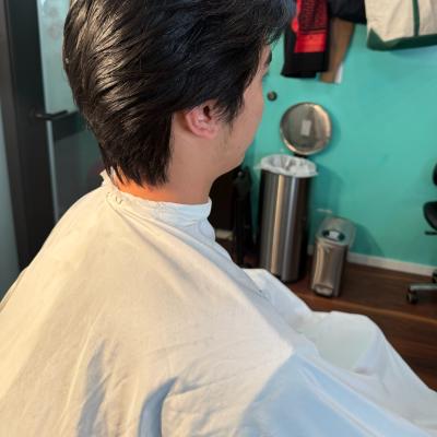 Asian man with a styled side-swept haircut, seated in barber chair wearing a cream cape, viewed from the side