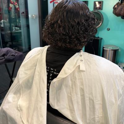 Person with voluminous curly dark hair, seated in barber chair wearing a white cape, viewed from behind
