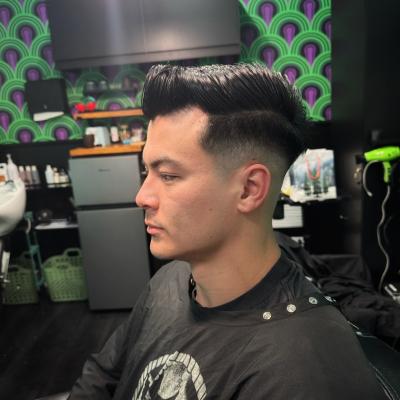 Man with a short dark taper fade haircut, seated in barber chair wearing a black graphic cape, profile view