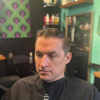Man with a short dark tapered fade, wearing a black graphic barber cape, viewed from the side
