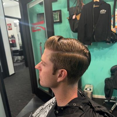Man with a short light-colored tapered haircut, wearing a black barber cape, profile view in barbershop