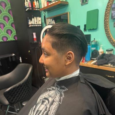 Man with short dark hair and a clean taper fade, seated in barber chair wearing a black cape, profile view