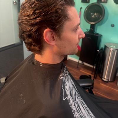Man with styled wavy medium-length brown hair swept back with volume, wearing a black patterned barber cape, profile view with teal barbershop wall in background