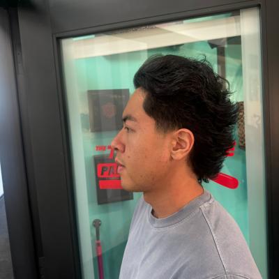 Man with short curly dark hair and a clean taper fade, wearing a light gray shirt, profile view with barbershop mirror and teal accents in background