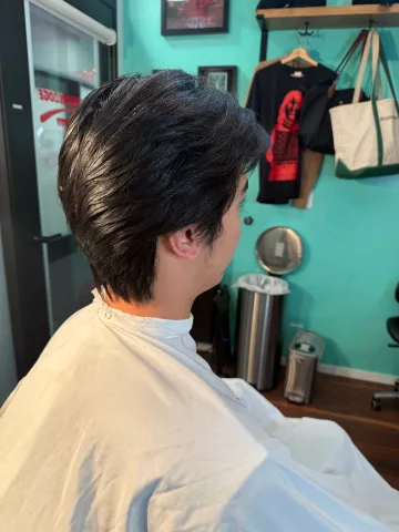 Asian man with a styled side-swept haircut, seated in barber chair wearing a cream cape, viewed from the side