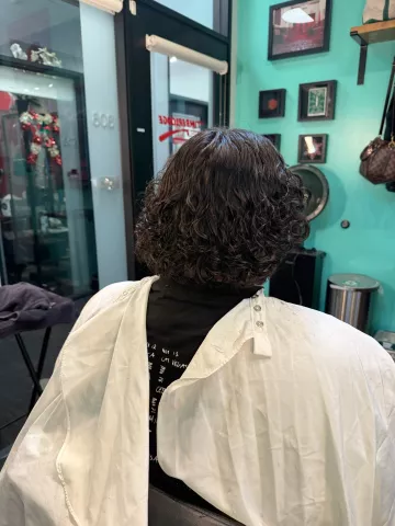 Person with voluminous curly dark hair, seated in barber chair wearing a white cape, viewed from behind