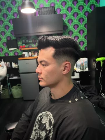 Man with a short dark taper fade haircut, seated in barber chair wearing a black graphic cape, profile view