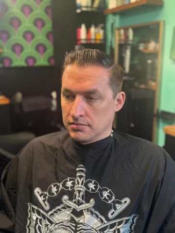 Man with a short dark tapered fade, wearing a black graphic barber cape, viewed from the side
