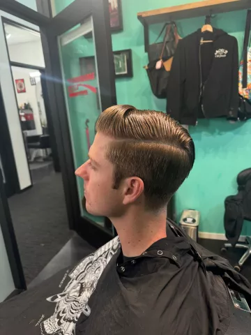 Man with a short light-colored tapered haircut, wearing a black barber cape, profile view in barbershop