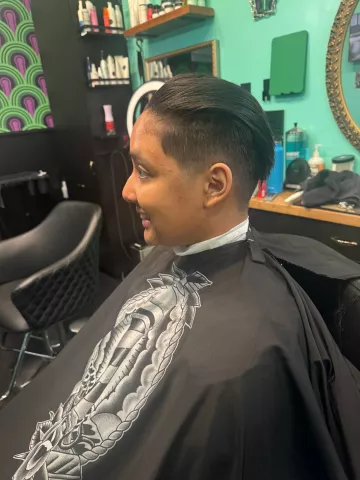 Man with short dark hair and a clean taper fade, seated in barber chair wearing a black cape, profile view