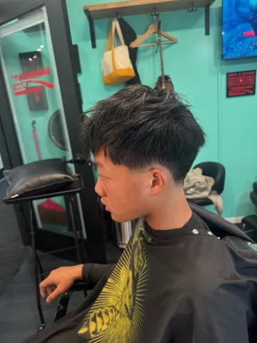Man with short dark hair and a skin fade, wearing a black barber cape, viewed from behind in barbershop