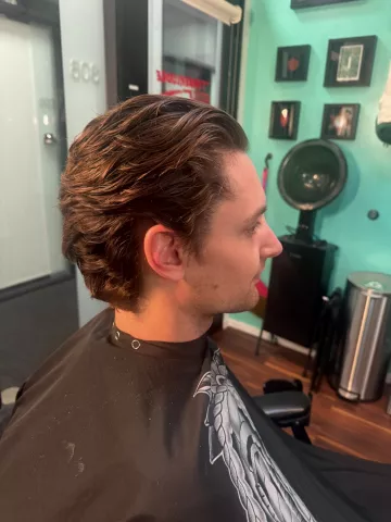 Man with styled wavy medium-length brown hair swept back with volume, wearing a black patterned barber cape, profile view with teal barbershop wall in background