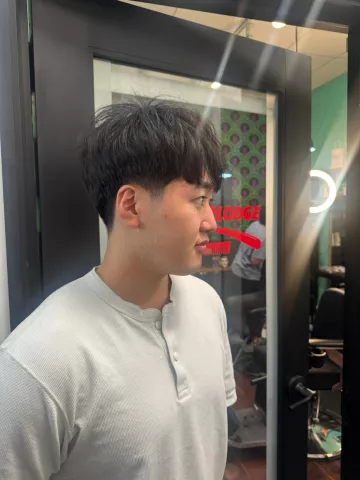 Asian man with a short blunt bowl cut with tapered sides, wearing a gray henley shirt, profile view reflected in barbershop mirror