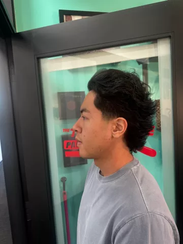 Man with short curly dark hair and a clean taper fade, wearing a light gray shirt, profile view with barbershop mirror and teal accents in background