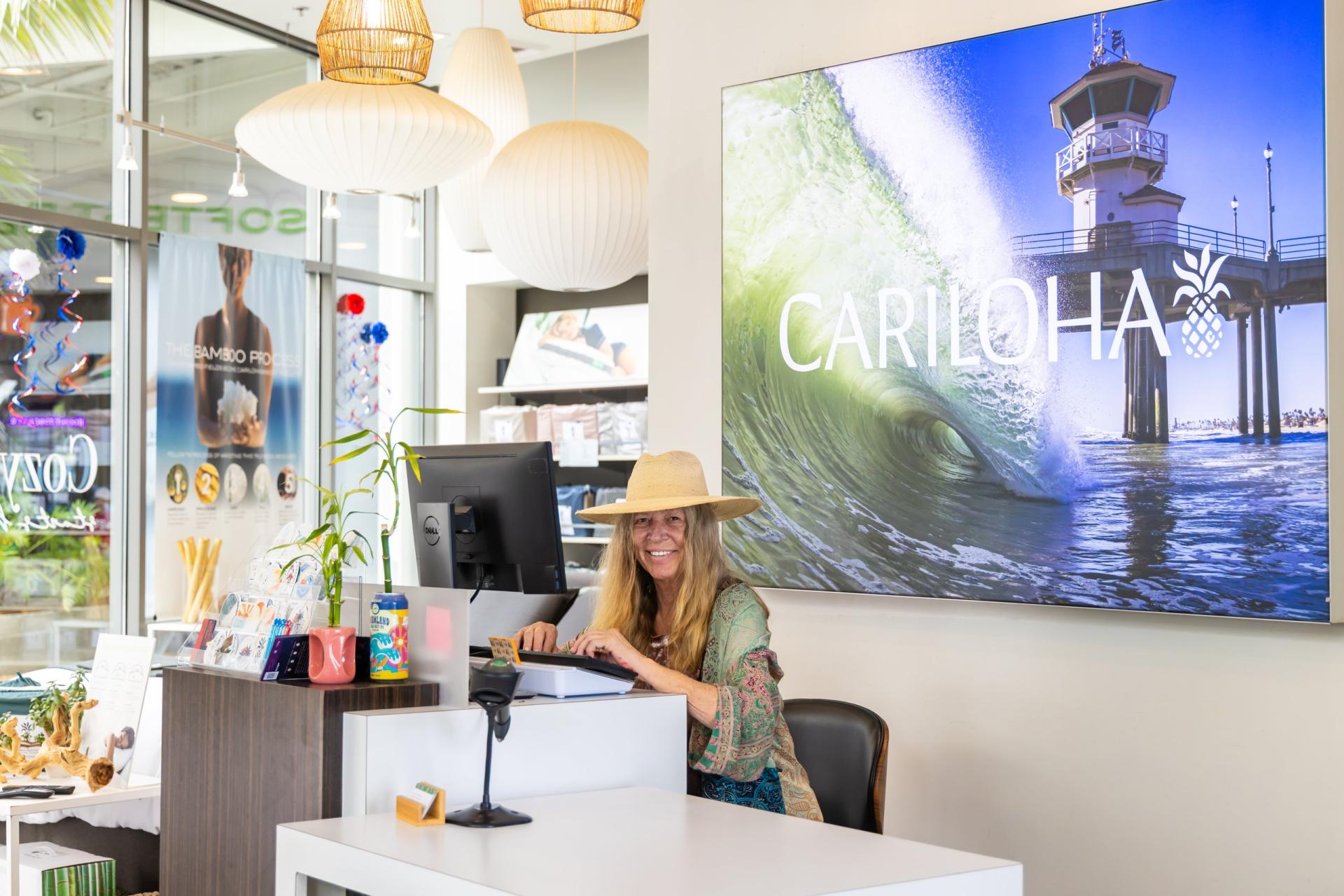 A woman in a straw hat smiles from the front desk at Cariloha, with a wall-sized photo of a wave and the Huntington Beach Pier behind her.