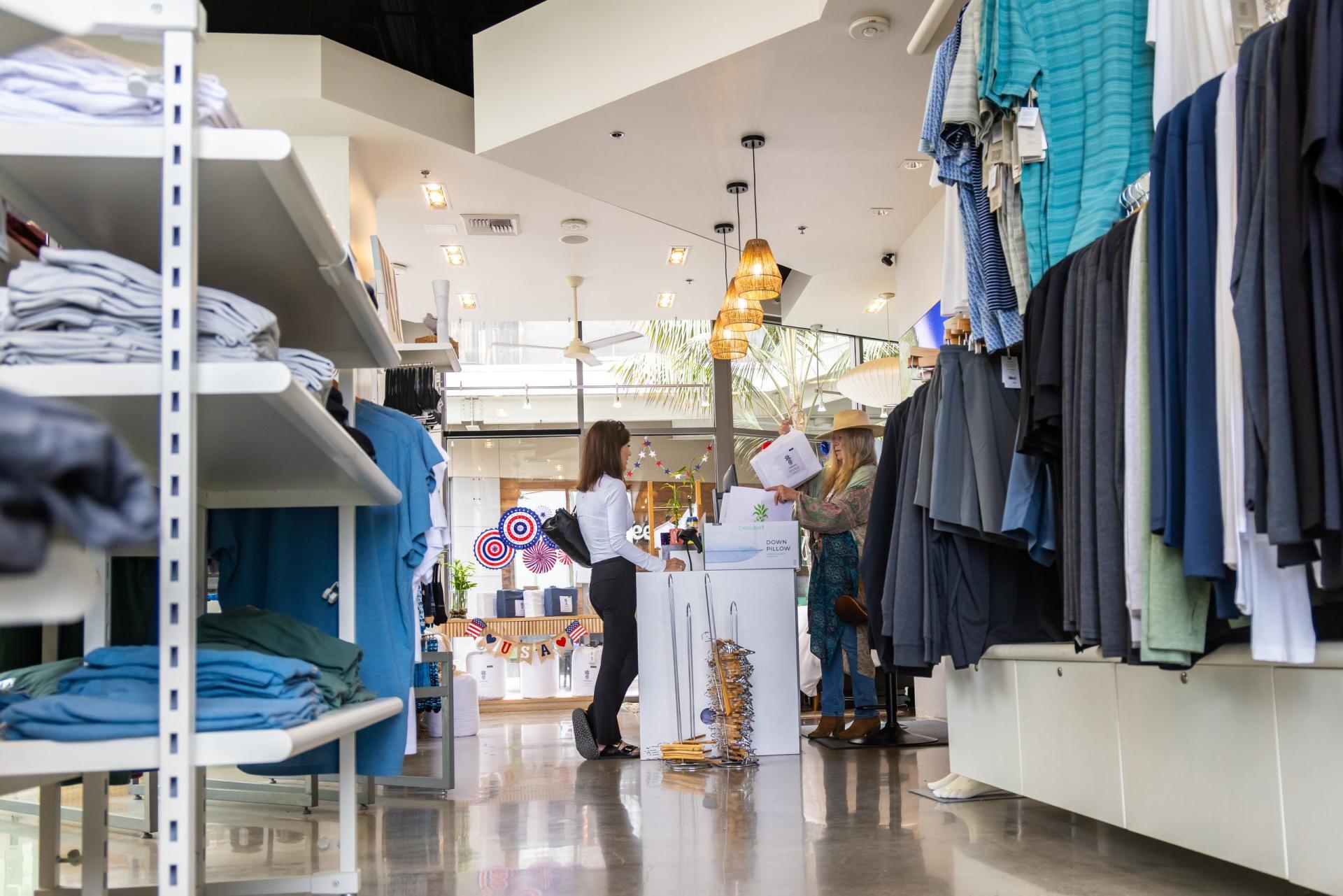 Folded shirts line one side of the aisle and hanging tees line the other, with two women at the counter by the front windows.